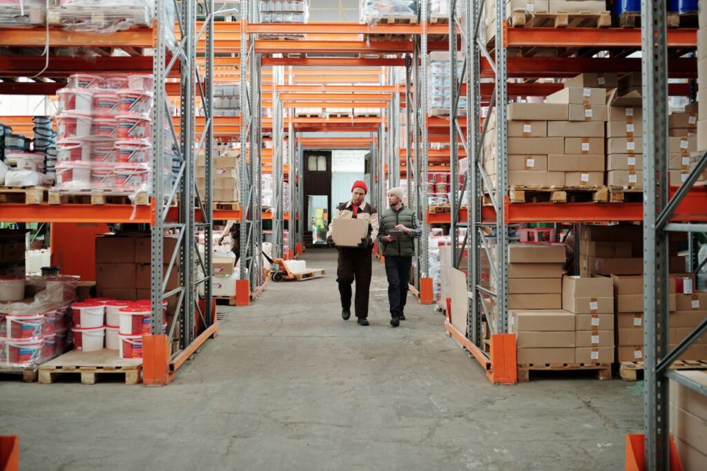 pexels photo 4487383 Two workers handle a package in a spacious warehouse surrounded by shelves stocked with boxes and products.